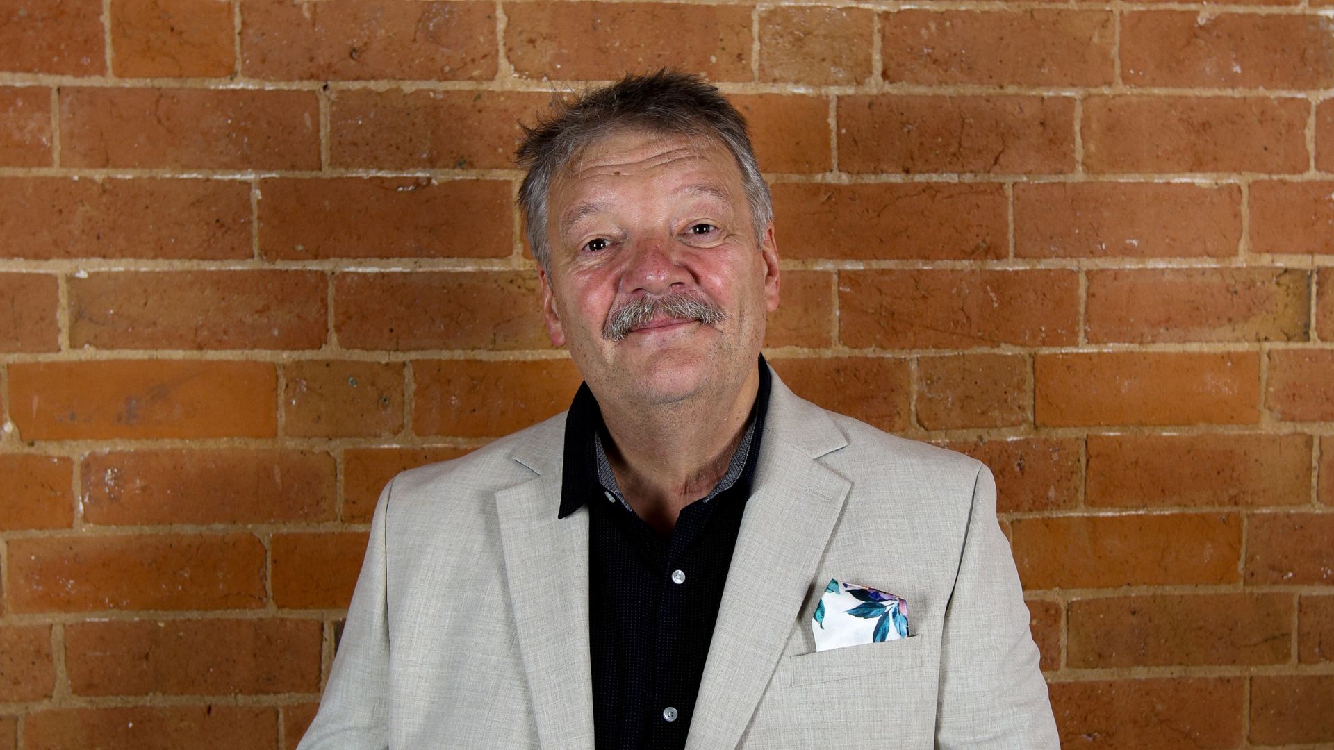 David-Cross David Cross standing against a brick wall in smart clothing, smiling at the camera and holding a tablet.